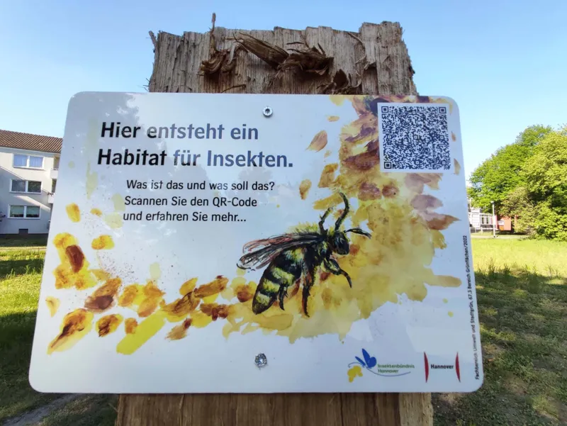 Ein Schild auf einer Bl&uuml;hwiese. Es enth&auml;lt Informationen und zeigt einen QR-Code.